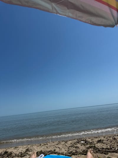 The image depicts a person's feet and lower legs resting on a sandy beach, with blue swimwear visible. The scene is set by the Black Sea, with calm, blue water stretching towards the horizon under a clear, bright blue sky. A portion of a colorful beach umbrella is visible at the top of the frame. The beach itself is covered in sand, with scattered debris such as dry reeds and seaweed. The location is identified as Bliznatsi, Bulgaria.