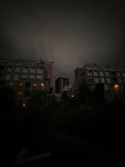The image captures a view of a city during a thunderstorm at night. In the foreground, silhouetted trees and foliage obscure the lower half of the frame. Above the trees, modern apartment buildings with visible lit windows dominate the mid-ground. One building, constructed of grey brick, is on the left, while another, also brick, is on the right. Between them, a taller, dark rectangular building stands in the background. Further in the background, a partially constructed orange tower with a red crane atop it is visible. The sky is dark and cloudy, with several streaks of lightning illuminating the upper portion of the scene. The overall atmosphere is one of a stormy urban night.