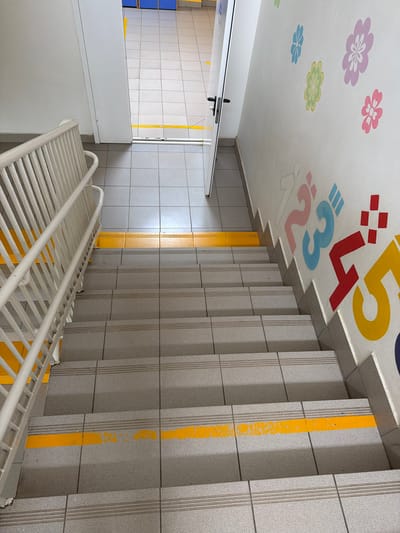 The image depicts a sterile, indoor staircase with gray tiled steps and a white railing. A bright yellow stripe runs across each step, serving as a safety marking. The walls are painted white and decorated with colorful floral patterns and large numbers in pink, blue, red, and yellow. A closed white door is visible at the top of the stairs, to the left of the decorated wall. Beyond the door, a hallway with tiled flooring and blue benches can be seen. The scene appears to be a public space, possibly a school or community center, located in Shcherbinka, Russia.