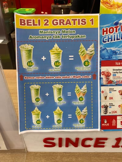 The image shows a promotional poster for a "Beli 2 Gratis 1" (Buy 2 Get 1 Free) offer for melon-based desserts. The poster is displayed in a shop located in Kecamatan Piyungan, Indonesia, at the coordinates -7.837 latitude and 110.474 longitude. The poster features three different dessert combinations, each consisting of two 16k (Indonesian Rupiah) drinks and one 8k melon ice cream. The drinks are labeled as "Milky Fruity Melon" and "Melon Kiwi Sundae." The ice cream is labeled as "Melon Ice Cream / Melon Mix Ice Cream." The poster also mentions "Semua varian dalam satu paket! Wajib coba!" (All variations in one package! Must try!). The poster is displayed in a shop with a wooden counter, and is likely intended to attract customers to purchase these desserts.