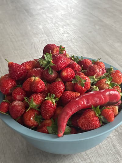 A light blue bowl is filled with a generous amount of fresh, red strawberries. A single, long, red chili pepper is nestled amongst the strawberries, its glossy skin reflecting the light. The bowl and its contents are placed on a light-colored, textured surface that appears to be wood or a similar material with a visible grain. The scene is shot from a slightly elevated perspective, looking down into the bowl. The lighting is natural and bright, suggesting it's daytime. The background is softly blurred, keeping the focus on the strawberries and the chili pepper. The overall impression is one of fresh produce, possibly prepared for a meal or for display. The setting appears to be indoors, given the lighting and the neutral background. There is no visible text in the image.