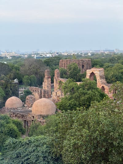 The image captures an elevated view of the Qutub Complex in Delhi, India. In the foreground, two large, weathered domes of ancient structures are visible, nestled amongst lush green trees and foliage. The domes appear to be made of stone or a similar material, exhibiting a slightly uneven surface. Behind these domes, the remnants of stone buildings rise, characterized by crumbling walls, arches, and pillars constructed from reddish-brown stone. Dense green trees and vegetation are interspersed throughout the complex, with some ancient trees featuring gnarled branches.

In the middle ground, beyond the immediate ruins, a modern, white, box-like structure is perched atop a metal platform supported by a dark, slender tower. Further in the distance, a sprawling cityscape unfolds under a hazy, overcast sky. Numerous buildings, ranging from low-rise structures to taller, modern edifices, form a dense urban landscape that extends to the horizon. The sky is a muted grey, suggesting either early morning, late evening, or overcast weather conditions. There are no visible people in the immediate foreground or middle ground of the image.