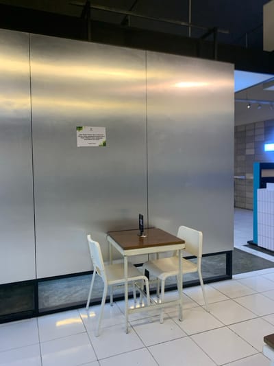 The image shows a small table and two white chairs in a cafe in Malang, Indonesia (coordinates -7.941, 112.608). The table is made of wood and has a brown tabletop. The chairs are made of metal and have white seats. The cafe has white tiled floors and a wall made of stainless steel. There is a sign on the wall with text in Indonesian. The sign is not visible enough to decipher what it says. The cafe is relatively empty, with no people present in the scene. The cafe is located in a building with black metal support beams. There is a window in the background that allows a glimpse of the outside world. The scene is lit by overhead lighting. There is also a small black box on the table. The table is centered in the image. The chairs are positioned on either side of the table. The floor is clean and white. The scene is mostly empty, with no people in the frame.