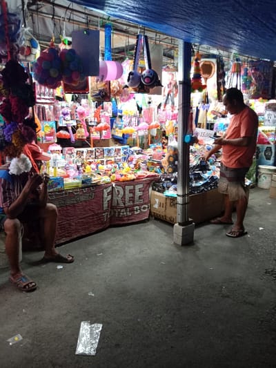 The image shows a street vendor stall in Kaliwungu, Indonesia, located at coordinates -6.962, 110.253. The stall is selling various toys and trinkets. A man in an orange shirt is standing at the counter, appearing to be browsing the selection of toys, while another man sits on the ground to the left of the counter. He is wearing a striped shirt and has his head down, possibly examining a small toy. The stall is covered by a blue tarp. There is a sign above the counter that says "FREE TOTAL BOX," with a brown, red, and white striped fabric on the counter below the sign. The ground in the image is a dark gray concrete.