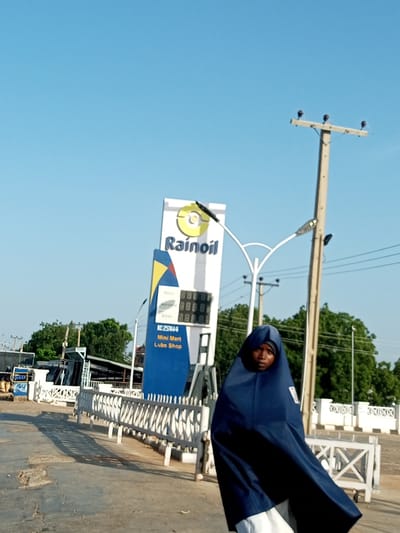 The image depicts a young person wearing a blue hijab and a white undergarment, standing outdoors in what appears to be a public area in Garko, Nigeria. Behind the person is a Rainoil gas station with a prominent white sign featuring the company logo and text indicating "Mini Mart Lube Shop." The gas station has a digital display showing numbers. Several utility poles with insulators are visible, along with trees and a white metal fence. The ground is paved and shows signs of wear. The sky is clear and blue. The scene suggests a typical day at a roadside service station in a Nigerian town.