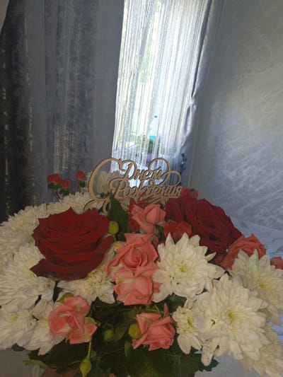 A bouquet of flowers, primarily white chrysanthemums and red and pink roses, is in the foreground. A wooden sign with Cyrillic text, likely "Happy Birthday," is nestled among the flowers. A window with sheer, vertical, string-like curtains allows light to filter into the room. Heavier gray curtains are on the left. The background is a textured, light gray wall. The location appears to be indoors, possibly in a home in Andreyevo, Russia.