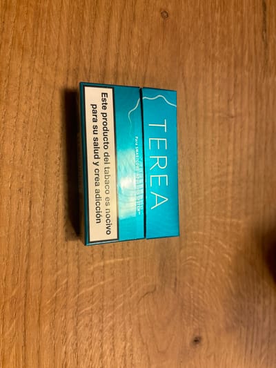 The image shows two packs of cigarettes, sitting on a wooden surface. The packs are blue and white, with the word "TEREA" printed in white letters on the right-hand pack. The left-hand pack has text printed in Spanish. The packs are likely located in Celerina/Schlarigna, Switzerland, based on the provided coordinates.