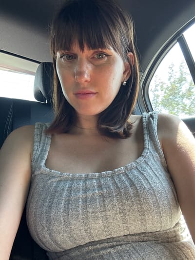 A woman with brown hair and bangs is seated in a car, likely near Pomezia, Italy, though specific landmarks are not visible through the car window. She is wearing a light gray, striped top. The car's interior is visible, including a black leather seat and a sunroof. The lighting suggests it is daytime.