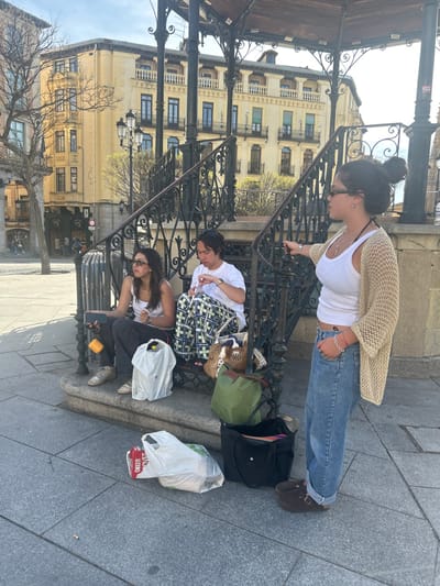 Three women stand and sit on a stone ledge in front of a wrought iron gazebo. The women are situated in front of a yellow building in the Plaza Mayor in Segovia, Spain (latitude: 40.95, longitude: -4.124). The woman on the right is wearing a white tank top, denim jeans, a beige crocheted sweater, and brown sandals. She is leaning against a black railing with her left arm, looking off to the left. The woman in the center is wearing a plaid shirt, white undershirt, and blue jeans. She is sitting with her left leg crossed and her hands on her lap. The woman on the left is wearing a black tank top, jeans, and black sneakers. She is sitting with her right leg crossed, holding a white bag in her lap, and looking down at a smartphone in her left hand. There are several bags and shopping bags on the stone ledge. The background includes a yellow building with black windows, an elaborate metal gazebo, and a large plaza. The sky is partly cloudy.