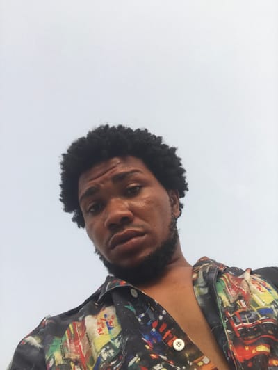 A person with dark skin, a black beard, and dark, curly hair fills the frame. They are wearing a colorful button-down shirt with abstract patterns. The photo is taken from a low angle, looking up at the person. The background appears to be a light grey or white, possibly the sky. The person's expression is serious and they appear to be tilting their head slightly.