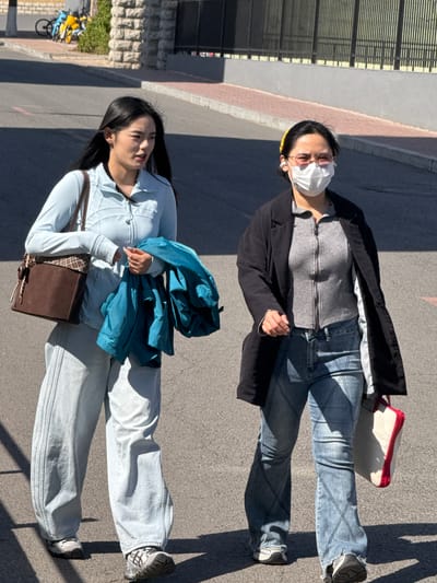 Two young women are standing on a paved surface outdoors. One is wearing a white face mask, a gray zip-up sweater, blue jeans, and a black jacket. She also has a yellow headband. The other woman wears a light blue zip-up jacket, light blue jeans, and carries a dark brown purse. A blue jacket is draped over her arm. The background shows a building with a brick base and barred windows. Bicycles are visible parked alongside the building. The location is Zhongtun, China.
