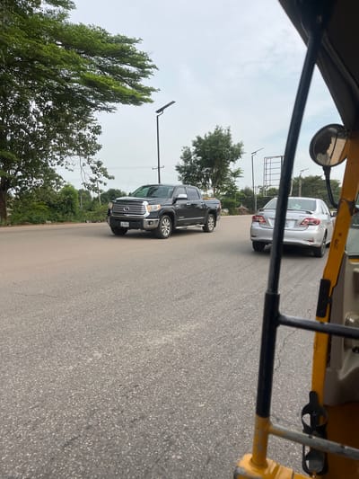 The image captures an outdoor street scene in Kuchingoro, Nigeria, taken from the interior of a vehicle, likely an auto-rickshaw, indicated by the visible yellow and black metal frame and a side-view mirror. The perspective looks out over a paved asphalt road beneath an overcast, bright sky.
In the midground, two vehicles are prominently featured. A silver Toyota Corolla sedan is seen from the rear, with the word "COROLLA" legible on its trunk. The rear left bumper of this car appears to be damaged or dented. Its license plate is partially visible, beginning with "ABJ 6..". Ahead of or beside the Corolla, a dark-colored pickup truck, identifiable by the "TOYOTA" badge on its front grille, is either parked or moving slowly. Its license plate, "ABJ 428WFA", is clearly visible.
The background features lush green trees lining the roadside, along with several utility poles or streetlights equipped with solar panels. The sky is uniformly bright but grey, suggesting an overcast daytime. The "ABJ" prefix on the vehicle license plates indicates the location is within Abuja, which is consistent with Kuchingoro being a district of the city. The scene depicts a common street environment with vehicles, but no people or specific activities are discernible.