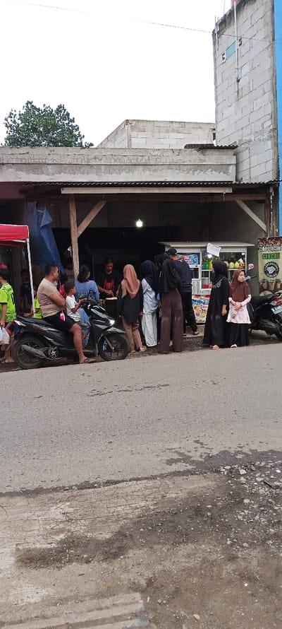 The image shows a street scene in Cangkuang, Indonesia (latitude: -7.055, longitude: 107.553). A group of people are gathered in front of a small shop, possibly a food stall. There are two motorcycles parked in front of the shop. Several people are wearing traditional clothing, and some are standing in line or talking to each other. A man wearing a blue shirt and brown shorts is seated on a motorcycle with a young boy beside him. A woman in a black shirt and black pants is standing in front of a food stall, possibly preparing food. The shop is enclosed by a concrete roof structure, and a grey concrete wall stretches beyond the shop. Several other concrete buildings are visible in the background. The street is unpaved and dusty.