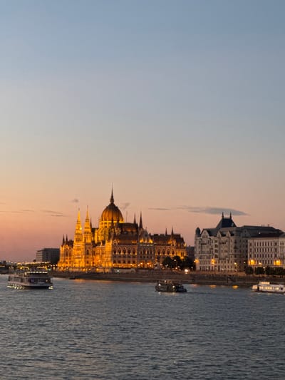 This media file depicts the Hungarian Parliament Building in Budapest at dusk. The iconic building, illuminated by warm lights, is situated on the bank of the Danube River. The river's surface is rippled, reflecting the lights of the building and the sky. Several boats are docked or moving along the river.
The sky displays a gradient of colors, transitioning from a soft blue at the top to hues of orange and pink near the horizon, indicating sunset or twilight. A few wispy clouds are visible against the sky. The architecture of the Parliament Building is intricate, with numerous spires and a prominent central dome. Adjacent buildings, also lit, line the riverbank.
The scene evokes a sense of peacefulness and grandeur. The time of day suggests a transition from day to night, with the artificial lights of the city beginning to dominate the natural light. There is no visible text in the image. The location cues, specifically the recognizable Parliament Building and the Danube River, confirm the setting as Budapest, Hungary.