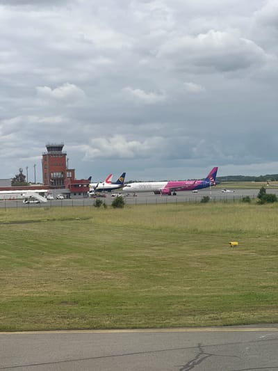 This image presents an aerial view of an airport tarmac and its surroundings, likely at Beauvais-Tillé Airport in Tillé, France. The scene is set under a partly cloudy sky during what appears to be daytime.

In the foreground, a large commercial aircraft, identifiable as a Wizz Air plane, dominates with its distinctive pink and purple livery. The text "WIZZ AIR" and "wizzair.com" are clearly visible along its fuselage. This aircraft is an Airbus A320 family model, and several ground support vehicles, including stairs, are positioned around it, suggesting active ground operations or passenger handling. Behind the Wizz Air plane, parts of at least two other commercial aircraft are visible, one with a blue and white tail featuring some red elements, and another predominantly white with a blue tail.

To the left of the parked aircraft stands a multi-story airport control tower, constructed primarily from brick, with its control cab visible at the top. Adjacent to the tarmac and control tower is a expanse of green grassy field, which separates the paved aircraft parking area from the distant tree line. A grey asphalt runway or taxiway, marked with yellow double lines, runs along the right side of the frame, showing some surface wear and cracks. Scattered yellow ground markers are visible in the grass. The sky above is filled with moderate cloud cover. The overall scene captures a typical, active airport environment.