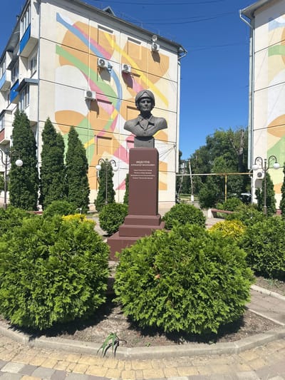 The image depicts a bust monument of a man in a military-style helmet and uniform, placed on a brown pedestal. The pedestal has text inscribed on it, but it is partially obscured by the bust. The monument is surrounded by manicured green bushes and cypress trees. In the background, there is a multi-story building with a colorful mural painted on its facade, featuring abstract geometric shapes and lines in various pastel colors. Two air conditioning units are visible on the building's exterior. The sky is clear and blue. The pavement in the foreground is made of interlocking bricks in yellow and grey. Streetlights with ornate designs and white globes are visible on the sides. The scene appears to be an outdoor public space in a city, likely a park or a square, in Armavir, Russia.