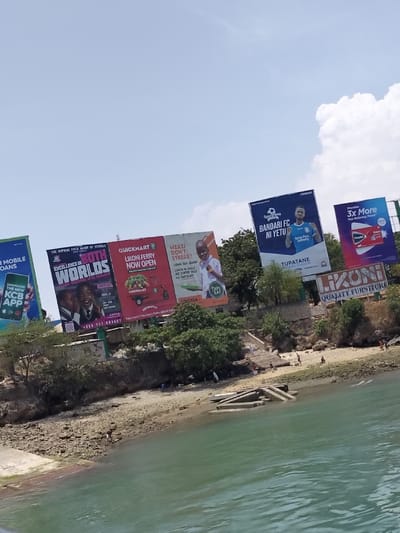 The image depicts a waterfront scene in Mkomani ward, Kenya, under a clear sky with scattered white clouds. The foreground features a body of greenish-blue water with slight surface ripples. Adjacent to the water is a rocky and sandy shoreline, characteristic of a beach.
On the shoreline, partially submerged concrete structures resembling pier remnants or foundations extend into the water on the right side. Multiple human figures are sparsely distributed along the beach, engaged in various, indistinct activities such as standing or walking.
Above the beach, a vegetated embankment or cliff rises, densely populated with green trees and shrubs. Prominently displayed along this elevated terrain are seven large commercial billboards. From left to right:
1. A partially visible green and blue billboard advertises "KCB Mobile Loans" and the "KCB App."
2. A red and white billboard promotes "The Supreme Field Group of Schools" with the slogan "Excellence in Both Worlds," featuring two school-aged children.
3. A red billboard for "Quickmart Likoni Ferry Now Open," displaying a red delivery tricycle (tuk-tuk) laden with produce.
4. A yellow and orange billboard with a young boy holding a tablet, advertising "Mzazi Don't Stress!!" and mentioning "Tapi Eat" and "Fontela."
5. A blue billboard for "SportPesa Bandari FC Ni Yetu," featuring a male football player in a blue jersey, associated with "Kenya Ports Authority."
6. A purple and blue billboard advertising "Pawa" with "3x More Pain Relieving Power," showing a product package.
7. A smaller billboard situated below the others on the right, displaying "LIKONI QUALITY FURNITURE."
A set of concrete stairs descends the embankment towards the beach on the right side of the image, with several individuals visible on or near these steps. Further human figures are present on the higher ground of the embankment among the vegetation.