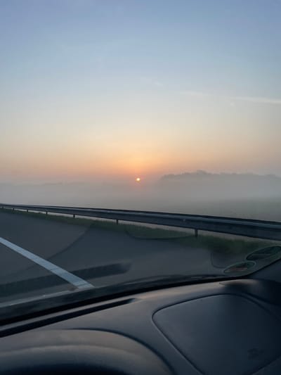 The image presents a view from the interior of a vehicle traveling on a road in Veldhoven, Netherlands, during sunrise or sunset. A bright, circular orange sun is visible low on the horizon, positioned slightly left of the center. A dense layer of white fog or mist blankets the landscape beneath a distant, dark green tree line, which is partially visible on the right. The sky above transitions in color from light blue at the top to pale orange and yellow closer to the horizon. In the mid-ground, an asphalt road with a distinct solid white edge line extends into the distance on the left. A silver metallic guardrail parallels the road on the right, separated from the pavement by a strip of green grass. The dark gray dashboard and the lower section of the vehicle's windshield occupy the immediate foreground. No individuals are visible.