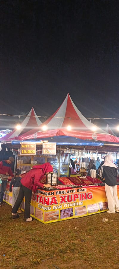 The image depicts an outdoor market stall at night, likely in Banding Agung, Indonesia, as indicated by the signage. Two red and white striped tents are prominent in the background. The foreground features a stall selling jewelry, identified by the banner "PUSTANA XUPING" and the text "PERHIASAN BERLAPIS EMAS" (gold-plated jewelry) and "XUPING DAN TITANIUM". Various pieces of jewelry are displayed on red fabric stands. Several people are present. A woman in a red hijab is examining the jewelry. A man in a blue and red jacket and a child in a dark shirt and grey pants are standing nearby. Another person in a black and white outfit and a white hijab is visible on the right. The ground is covered in grass and dirt. String lights are visible above the stall, illuminating the scene.
