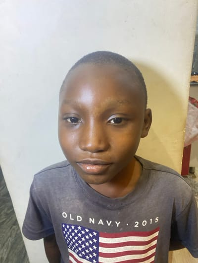 A close-up, slightly tilted shot features a young boy with short, dark hair. He has smooth, dark skin and dark eyes, looking directly at the camera with a gentle expression. He's wearing a gray t-shirt featuring a stylized American flag design along with white lettering that reads "OLD NAVY. 2015". The background consists of a plain, light-colored wall.