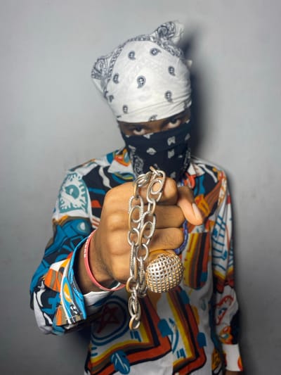 An individual wearing a colorful patterned shirt, a patchwork bandana, and jewelry is shown. A fist clenching a silver chain and a large, decorative silver ring is at the forefront. The sleeve of the shirt has vibrant geometric shapes and symbols. The bandana features sections of varying colors and patterns, including paisley and solid black. The person's skin tone is brown. The background appears to be a plain gray or white surface.