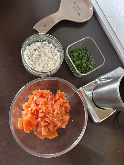 A counter in what appears to be a kitchen setting in Aprilia, Italy features a preparation scene for a meal. A clear glass bowl filled with diced salmon pieces is prominent. Next to it, another clear glass bowl contains finely diced white onion. A small, square glass container holds chopped green herbs, possibly parsley or cilantro. A light-colored, ceramic spoon rest with Italian text ("Ma Che Bontà!") is also visible. In the upper right quadrant is what appears to be a silver kitchen appliance, possibly a blender or food processor. The countertop is a dark color, and the background includes a white tiled wall. No people are visible. The focus is on the preparation of ingredients.