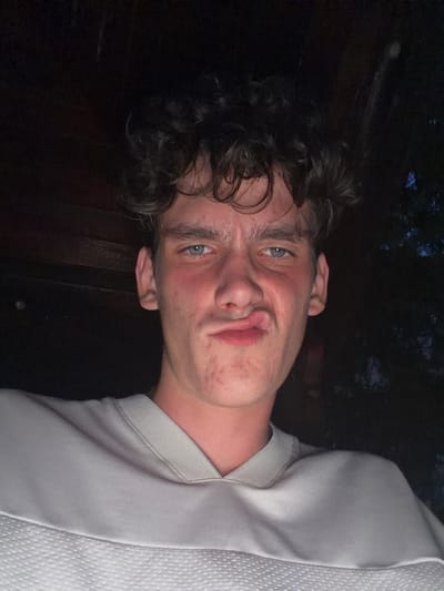 A young male, approximately late teens to early twenties, is depicted in a close-up portrait, located in Dupnitsa, Bulgaria. He has a fair complexion with visible skin texture and some facial blemishes, dark curly hair, and light blue eyes. His lips are deliberately puckered and twisted to the left, forming a humorous or exaggerated expression, while his brows are slightly furrowed. He is wearing a light grey long-sleeved top featuring a ribbed collar and a textured fabric panel across the chest. The lighting is dim, suggesting an evening or nighttime setting. The background is predominantly dark, with a dark wooden structure visible on the left side, and faint, blurred dark outlines, possibly foliage or trees, on the right, indicating an outdoor or semi-outdoor environment.