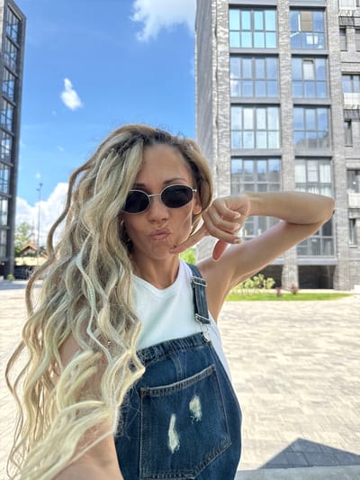 A woman with blonde dreadlocks and sunglasses is taking a selfie outdoors in front of a modern brick building with large windows. She is wearing a white top and denim overalls. She is making a kissing face and gesturing with her hand. The background shows a clear blue sky with some clouds, the building, and a paved area. The location is identified as Rusanovo, Russia.