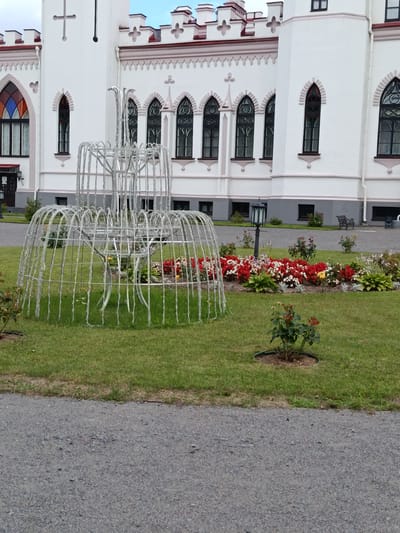 The image shows the Kosava Castle in Kosava, Belarus. The castle is a large, white building with a distinctive architectural style, featuring pointed arches, crenellations, and stained-glass windows. In front of the castle, there is a manicured lawn with a decorative metal structure that resembles a fountain. A flower bed filled with red and white flowers is also visible, along with a lamppost and a park bench. The scene appears to be set on a cloudy day, with the sky visible in the background. The image is captured from a ground-level perspective, looking towards the castle.