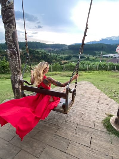 A woman with long blonde hair and tattoos on her arms is sitting on a swing. She is wearing a long, flowing red dress. The swing is made of metal and wood, and it is suspended by chains. The woman is holding onto the chains with her hands. She is looking off to the side, and her expression is serene.
The background of the image is a lush green landscape with rolling hills. There are some trees and a cloudy sky. The ground is covered in paving stones. The image appears to have been taken during the daytime. The weather is overcast.
The woman's red dress is vibrant and contrasts with the green landscape. The tattoos on her arms add an interesting detail to her appearance. The swing is rustic and complements the natural setting. The overall impression is one of peacefulness and natural beauty.