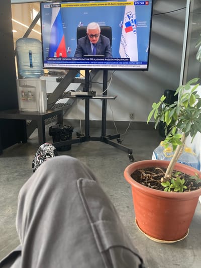 The image captures a casual moment in an office environment, with a television screen displaying a news broadcast from Russia 24. The broadcast features a man in a suit and glasses speaking, with the Russian flag and the logo of the Russian Union of Industrialists and Entrepreneurs visible in the background. Below the broadcast, a text overlay reads "Совещание с членами Правительства РФ в режиме видеоконференции" (Meeting with members of the Government of the Russian Federation in video conference mode). The foreground is dominated by the legs of a person wearing gray sweatpants and an athletic shoe. To the left of the television, a water cooler with a large bottle of water is situated on a black table. Nearby, a trash can and a potted plant with green leaves in a terracotta pot add to the office setting. The overall scene suggests a workspace in Sarapul, Russia, where an individual is taking a break to watch the news.