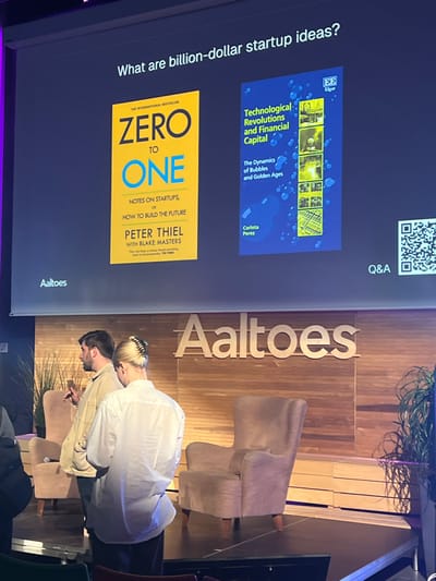 The image depicts an indoor presentation venue, identified by the prominent "Aaltoes" logo on a wooden panel wall behind the stage, indicating Aalto University in Espoo, Finland. A large projection screen displays the title "What are billion-dollar startup ideas?" centrally. Below this title, two book covers are shown: "Zero to One" by Peter Thiel (left, yellow) and "Technological Revolutions and Financial Capital" by Carlota Perez (right, blue). The screen also displays "Aaltoes" on the bottom left and "Q&A" with a QR code on the bottom right.

On the stage, two individuals are present. A male, wearing a light-colored shirt and possessing a beard, stands to the left, holding a microphone in his right hand and gesturing with his left. He faces left, seemingly interacting with an unseen audience or another individual. A female, with her back mostly to the camera, stands to the right of the male, wearing a white collared shirt and dark pants. Her blonde hair is secured with a large claw clip. The stage is dark-surfaced and features two light-colored upholstered armchairs, one visible on the left and one on the right. Potted green plants are positioned at the edges of the stage.
