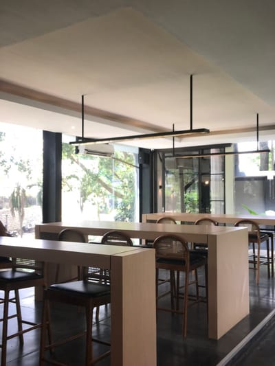 The image shows the interior of a modern restaurant located at coordinates -7.98 latitude and 112.617 longitude, placing it in Malang, Indonesia. The restaurant has a white ceiling with two long, black, horizontal light fixtures hanging from it. The floor is dark grey tile and the walls are painted white. Three long, light-brown wooden tables line the far wall. Each table is set with four black leather-backed chairs with woven cane seats. The tables are placed near a large window that looks out onto a green, leafy outdoor area. There are no people visible in the image. The space appears to be a restaurant or cafe. The tables and chairs suggest that it is a place for dining. The modern design and the bright lighting suggest that it is a contemporary establishment. The cleanliness and well-kept appearance suggest that it is well-maintained and popular.