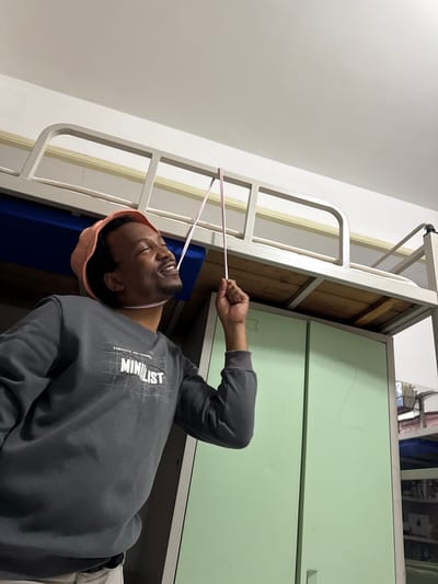 In the Daxing District of China, a man with dark skin and a light peach bucket hat is positioned in the foreground. He is wearing a dark grey sweatshirt with the word "MINILIST" in white lettering. His eyes are closed, and he is looking upwards with a smile. His right hand is raised, holding two thin pink sticks. He appears to be in a dorm room, with a bunk bed above him and light green cabinets to his right. The ceiling is white, and the overall lighting is soft and indoor.