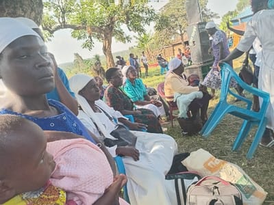 This image depicts an outdoor gathering, primarily of women and children, in a rural or semi-rural setting, consistent with Burundu, Kenya. The scene is illuminated by bright daylight, suggesting midday or early afternoon.
In the foreground, a woman wearing a blue top and a white head covering holds a baby wrapped in a pink blanket. Her expression appears serious or contemplative. She is seated on a blue plastic chair, as are several other women positioned behind and to her right. Many of these women also wear white head coverings and are dressed in various colored garments, including patterned dresses and solid blue or green tops. One woman further back in a yellow top is seen holding a child.
On the right side of the frame, a large black speaker with the text "ROYAL SOUND" and a partial phone number "075...19194" is prominent, suggesting music or announcements are part of the event. A woman in a purple patterned dress is standing near the speaker, while another person in a white top appears to be moving a blue plastic chair. Various bags, including a grey duffel bag and a white sack with "FRESH" printed in green, are visible on the ground in the lower right foreground.
The gathering takes place on a grassy area, shaded by large trees with green leaves. In the distant background, what appear to be simple village structures or huts can be seen. The overall impression is of a community or social event, possibly with a religious or cultural dimension, given the attire of many participants.