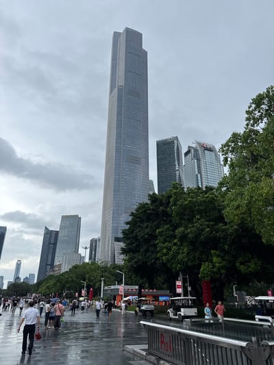 The image captures a street scene in Tianhe District, China, under an overcast sky, with wet pavement indicating recent rain. A very tall, modern skyscraper dominates the background, with other contemporary buildings of varying heights also visible. Lush green trees line the street, interspersed with the urban architecture. A paved walkway is filled with a diverse crowd of people, many carrying umbrellas or dressed in casual attire, suggesting a public area with pedestrian traffic. Several small, open-air electric vehicles, likely for tours or transportation, are present on the right side of the image. The overall impression is of a bustling urban environment in a city center.