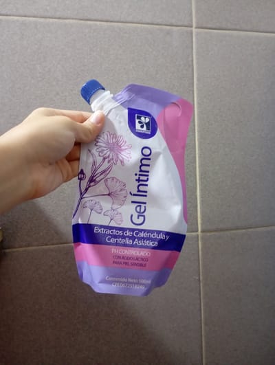 A person's hand is holding a purple and white pouch of "Gel Intimo" in front of a tiled wall. The pouch has floral illustrations and text in Spanish. The text on the pouch indicates it contains extracts of calendula and centella asiatica, is pH controlled, with lactic acid for sensitive skin, and has a net content of 500ml. The tiles on the wall are a neutral gray color and appear to be in a bathroom or similar environment. There is no specific visual information in the image that would allow for the determination of its location within the city of La Tagua, Venezuela.