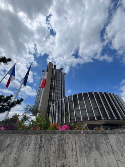 This media file captures a low-angle view of a modern building with a glass facade, set against a bright blue sky dotted with white clouds. In the foreground, three flags are visible on flagpoles: the European Union flag, a French flag, and another French flag. The building itself appears to be tall and has a distinctive, ribbed exterior.
In the middle ground, there is a section of the building that appears to be covered in solar panels. Lush greenery and vibrant pink and orange flowers spill over from planters situated along the building's edge, adding a touch of color to the urban landscape. A concrete wall is prominently featured on the right side of the frame, providing a textural contrast to the sleekness of the building.
The image suggests a clear day, likely daytime, given the brightness of the sky and the strong shadows. The presence of the European Union and French flags indicates the location is likely in France, and the provided context confirms it is Créteil, France. No people are visible in the frame. The overall impression is one of modernity, civic presence, and a touch of natural beauty incorporated into an urban setting.