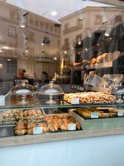 The image shows a bakery storefront window display in Segovia, Spain (latitude: 40.947, longitude: -4.119). The display case features a variety of baked goods, including croissants, pastries, and bread. There are two glass dome covers, one empty and one partially filled with pastries. The bottom shelf is filled with several types of pastries, with handwritten signs denoting their names and prices. Several more shelves are stacked with other pastries, with the topmost shelf featuring bread. The display is lit from above and the window reflects the cityscape behind it, including buildings with balconies and a person in the distance. The reflection is slightly blurred, showing that it is viewed through a glass surface. The bakery is open and inviting.