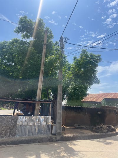The image shows an outdoor scene, likely in Jimeta, Nigeria, on a sunny day with scattered clouds. The perspective is slightly tilted, looking upwards towards a utility pole adorned with electrical wires and transformers. A large tree with lush green foliage dominates the central part of the frame, its branches reaching towards the sky. In the background, a building with a rusty, corrugated metal roof is visible, topped with barbed wire. The building's wall appears to be made of concrete and has some staining. To the right, a concrete pathway or road runs alongside the building, casting shadows from unseen objects. Further to the right, part of another building's wall, also made of concrete, is visible. The lower part of the image shows some corrugated metal sheeting attached to a stone wall, possibly as a repair or modification to a structure. The overall impression is one of an urban or semi-urban environment, with a mix of natural elements and man-made structures. There are no people or explicit activities depicted. The bright sunlight suggests it is daytime, likely late morning or early afternoon.