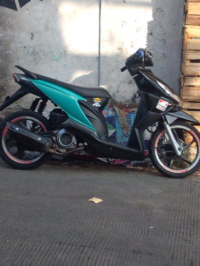 A black scooter with teal accents is parked on a paved surface in front of a concrete wall. The scooter has a sticker on the front fender with the Honda logo and another sticker on the seat depicting a Jolly Roger symbol. The rear of the scooter features a black exhaust pipe and a rimmed wheel with red accents. Wooden crates are stacked to the right of the scooter, and torn paper or posters are adhered to the concrete wall behind it. The ground is marked with parallel lines. The scene is illuminated by sunlight casting shadows.