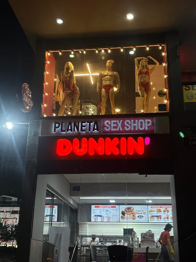 A storefront displays a jarring juxtaposition. On one side, mannequins in lingerie pose in a window labeled "Planeta Sex Shop". Next to this, the bright orange "Dunkin'" logo shines above the entrance to a Dunkin' restaurant. The contrast highlights the strange combination of services: explicit adult goods and a family-friendly donut chain in the same building or very close proximity. Street signs and a partial menu board for Dunkin' are also visible.