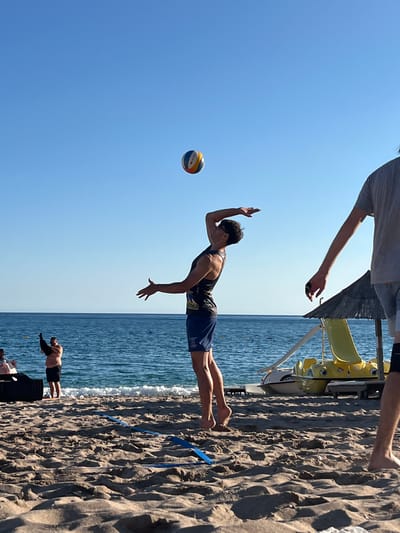 A beach volleyball game is in progress on a sandy beach. A man in a black tank top and blue shorts is in mid-air, jumping to hit a volleyball, which is visible in the sky above him. Another person, partially visible on the right side of the frame, appears to be his teammate or opponent. In the background, the blue sea stretches out to the horizon, with gentle waves lapping at the shore. Further back, a man in black shorts and no shirt is standing near a black object, possibly a beach bag or chair. To the right of the volleyball players, there is a yellow waterslide and a straw umbrella. The sky is clear and blue. The scene is likely in Sutomore, Montenegro, given the context provided.