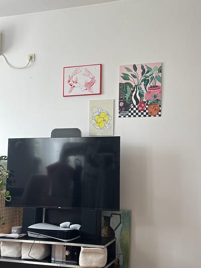 The image shows a living room with a large black television mounted on a stand. Above the television, three framed pictures hang on a white wall. The leftmost picture features two red koi fish. To its right is a smaller, yellow-toned picture with lemons. The largest picture, on the far right, depicts various potted plants and vases against a pink background with a checkered pattern. A black electrical outlet with a white cord is visible on the wall to the left of the pictures. Below the television, on a black shelf unit, are a gaming console, controllers, a digital clock displaying "2:42", and decorative baskets. To the right of the shelf, a vertical painting with a muted color palette featuring two vases is partially visible. The overall scene suggests a contemporary home interior. The location is not discernible from the image itself.