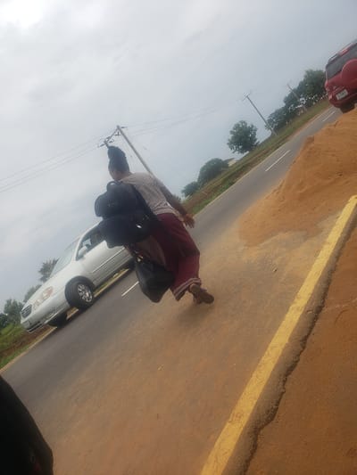 A woman, seen from behind, walks along a paved road in Gumel, Nigeria. She wears a patterned shirt and a maroon skirt, and carries a black backpack and a black plastic bag. A silver sedan is parked on the left side of the road, and a red SUV is visible further down the road on the right. The sky is overcast, and trees and power lines are visible in the background. The ground is dusty and brown, with a yellow line painted on the road.