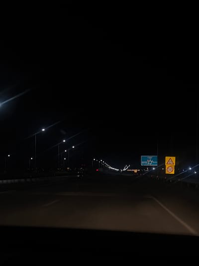 The image depicts a nighttime scene on a highway. Multiple streetlights illuminate the road, casting long shadows. In the distance, signs are visible, including a blue directional sign with a circular intersection symbol and text, and a yellow triangular sign with a red border and a speed limit sign showing "50". The road appears to be asphalt and has lane markings. There are no people or vehicles clearly visible in the foreground, though some faint lights in the distance might indicate other traffic. The environment is dark, with the sky being entirely black. The scene is likely on a highway leading into or out of Asenovgrad, Bulgaria, given the presence of road signs.