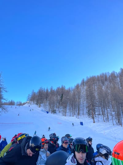 The image captures a vibrant winter scene at a ski resort in Courmayeur, Italy, with a clear blue sky overhead. The perspective is from the side of a ski slope, looking down towards the course and across to a forested mountainside.

In the foreground, a group of people, likely skiers or spectators, are clustered together. Most are wearing ski helmets and goggles, suggesting they are prepared for or participating in skiing activities. One person in a black helmet and green-tinted goggles looks directly towards the viewer. Visible helmet brands include "SPY," "POC," "BRIKO," and "O'NEILL." Part of a bright orange inflatable object, possibly a safety barrier or marker, is visible at the very edge of the frame.

The midground features a wide, snow-covered ski run. Several skiers can be seen making their way down the slope, some appearing to navigate around blue and red gates, indicating a slalom or giant slalom course. This suggests an organized event, such as a race or training session. The pristine snow of the slope contrasts with the darker line of trees in the background.

The background is dominated by a dense forest of bare, deciduous trees, likely larch, covering the mountain slopes. The trees are brown and leafless, typical of a winter alpine landscape. Above the forest, the sky is a brilliant and cloudless blue, indicating excellent weather conditions for skiing. The overall impression is one of a busy, active day at a scenic ski destination.