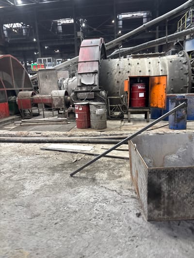 The image depicts the interior of an industrial facility, potentially a factory or plant, located in Mirkovo, Bulgaria. A large, cylindrical machine with numerous bolts and pipes dominates the scene. Several red barrels labeled "Mobil" are visible, indicating the presence of lubricating oil. Metal structures, including platforms, pipes, and beams, are scattered throughout the area. The floor is concrete and shows signs of wear and grime. A metal box with an open lid sits on the right side of the image. The overall impression is one of a functional, but somewhat cluttered and aging, industrial setting.