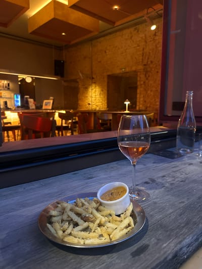 The image depicts a close-up view of a wooden table on which a plate of french fries with a dipping sauce, a stemmed wine glass filled with rosé wine, and a bottle of water are arranged. In the background, through a glass window, the interior of a restaurant is visible. The restaurant features exposed brick walls, wooden ceiling panels with integrated lighting, and tables with chairs. A bar area with bottles is also discernible. The overall ambiance suggests an evening setting, possibly in a restaurant or bar. The specific location in Vilnius, Lithuania, is not discernible from the image content.