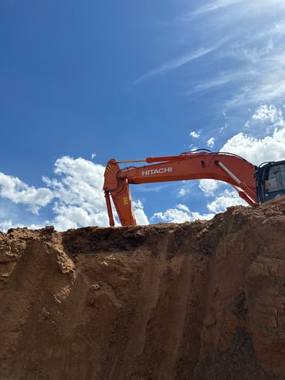 An orange Hitachi excavator is positioned on top of a mound of dirt under a bright blue sky with scattered white clouds. The excavator's arm is extended, and the brand name "HITACHI" is visible on the arm. The foreground of the image consists of a steep, uneven dirt embankment, likely the result of excavation. The scene appears to be a construction site, possibly in Astana, Kazakhstan, given the context provided. There are no people visible in the image.
